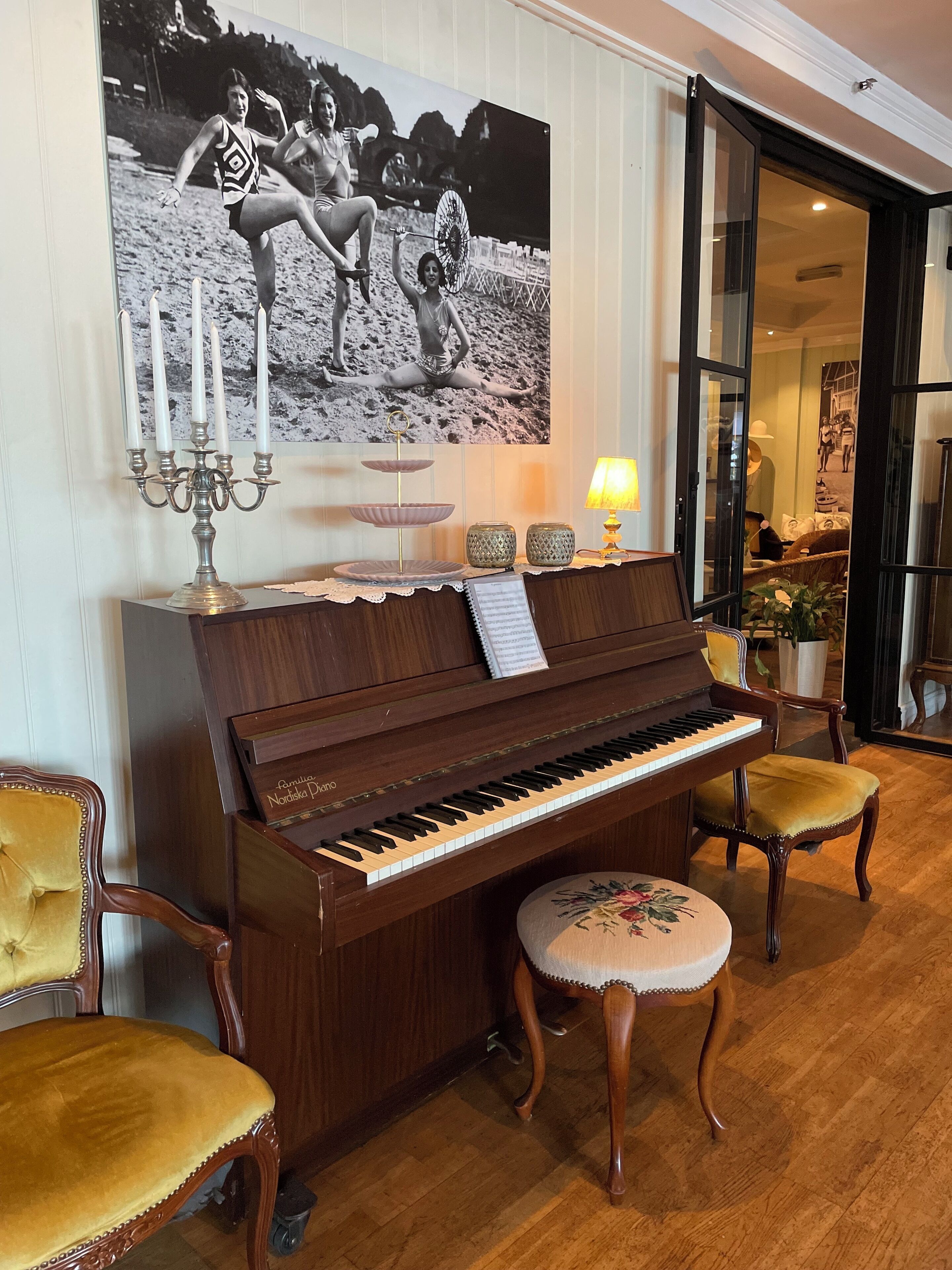 A grand piano in a well-lit room with a large black-and-white photograph on the wall, a candelabra, and a small table with a lamp.