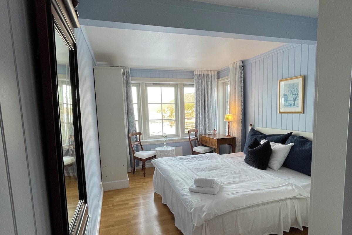 Un dormitorio con una cama grande, un escritorio y una silla. Se ve una ventana con cortinas y un cuadro enmarcado en la pared.