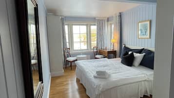A bedroom with a large bed, a desk, and a chair. There is a window with curtains and a framed picture on the wall.
