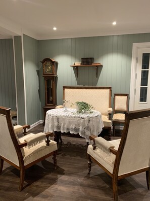 A dining area with a round table covered by a lace tablecloth, surrounded by four upholstered chairs with wooden frames. A wooden clock and a shelf with books are visible on the wall. - Badehotellet (Asker)