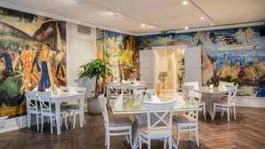 A dining area with white chairs and tables, a mural on the wall, and a potted plant.