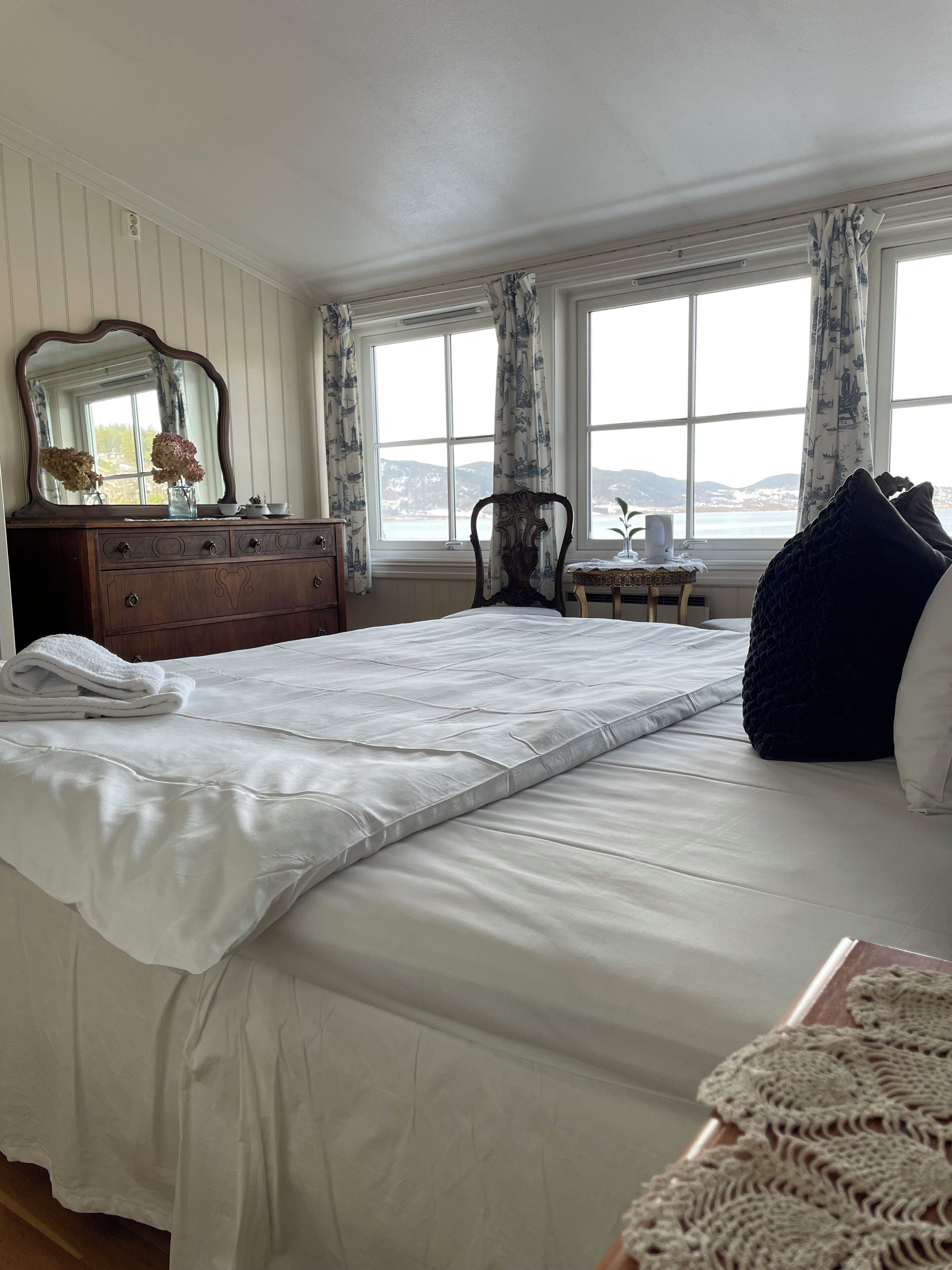 A bedroom with a large bed, a dresser with a mirror, a window with curtains, and a view of the outdoors.