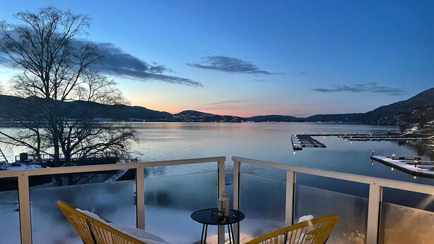 A balcony with two yellow chairs and a small table, overlooking a calm lake with a dock and mountains in the background.