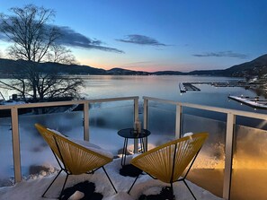 A balcony with two yellow chairs and a small table, overlooking a calm lake with a dock and mountains in the background. - Badehotellet (Asker)