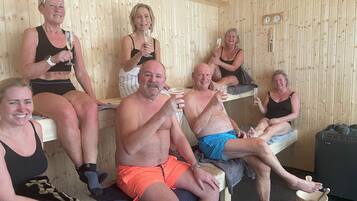 A wooden sauna with people sitting on benches, holding drinks, and a wooden bucket on the floor.