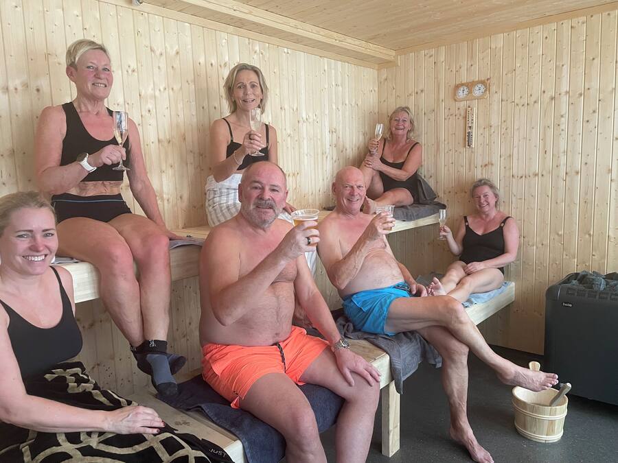 A wooden sauna with people sitting on benches, holding drinks, and a wooden bucket on the floor.