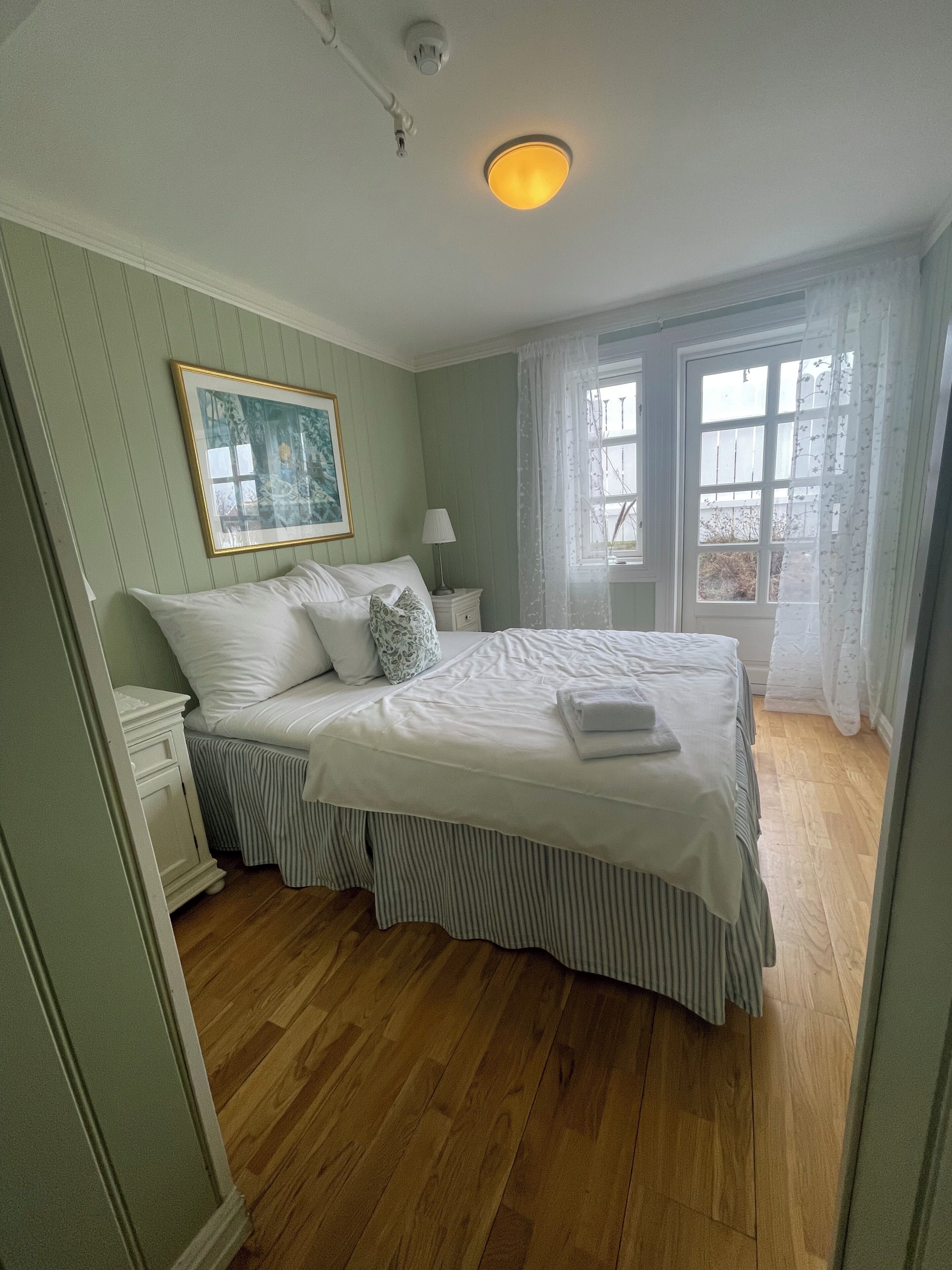 A bedroom with a bed, a nightstand, a lamp, a painting, and a window with curtains.