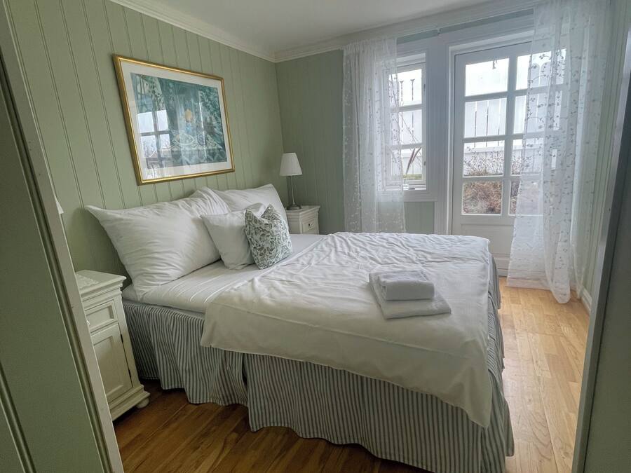 A bedroom with a bed, a nightstand, a lamp, a painting, and a window with curtains.