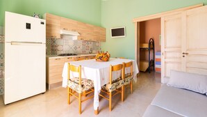A compact kitchen with a refrigerator, stove, and wooden cabinets. There is a dining table with chairs and a small TV on the wall. - 2038 Villetta Gloria (Alliste)