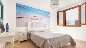 Ett sovrum med en stor väggmålning som föreställer en strand, en vit säng, ett skrivbord med en vas och ett fönster med träramar.