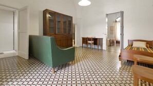 Une pièce avec un sol à motifs, une armoire en bois, un fauteuil vert, un lit recouvert d’une courtepointe colorée et un luminaire suspendu.