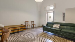 Une pièce avec un sol à motifs, un canapé vert, deux chaises en bois et un lit recouvert d’une couverture à rayures.