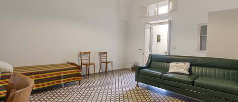 A room with a patterned floor, a green sofa, two wooden chairs, and a bed with a striped blanket.