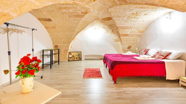Un dormitorio con techo de piedra, cama con colcha roja y un florero con flores rojas sobre una mesa.