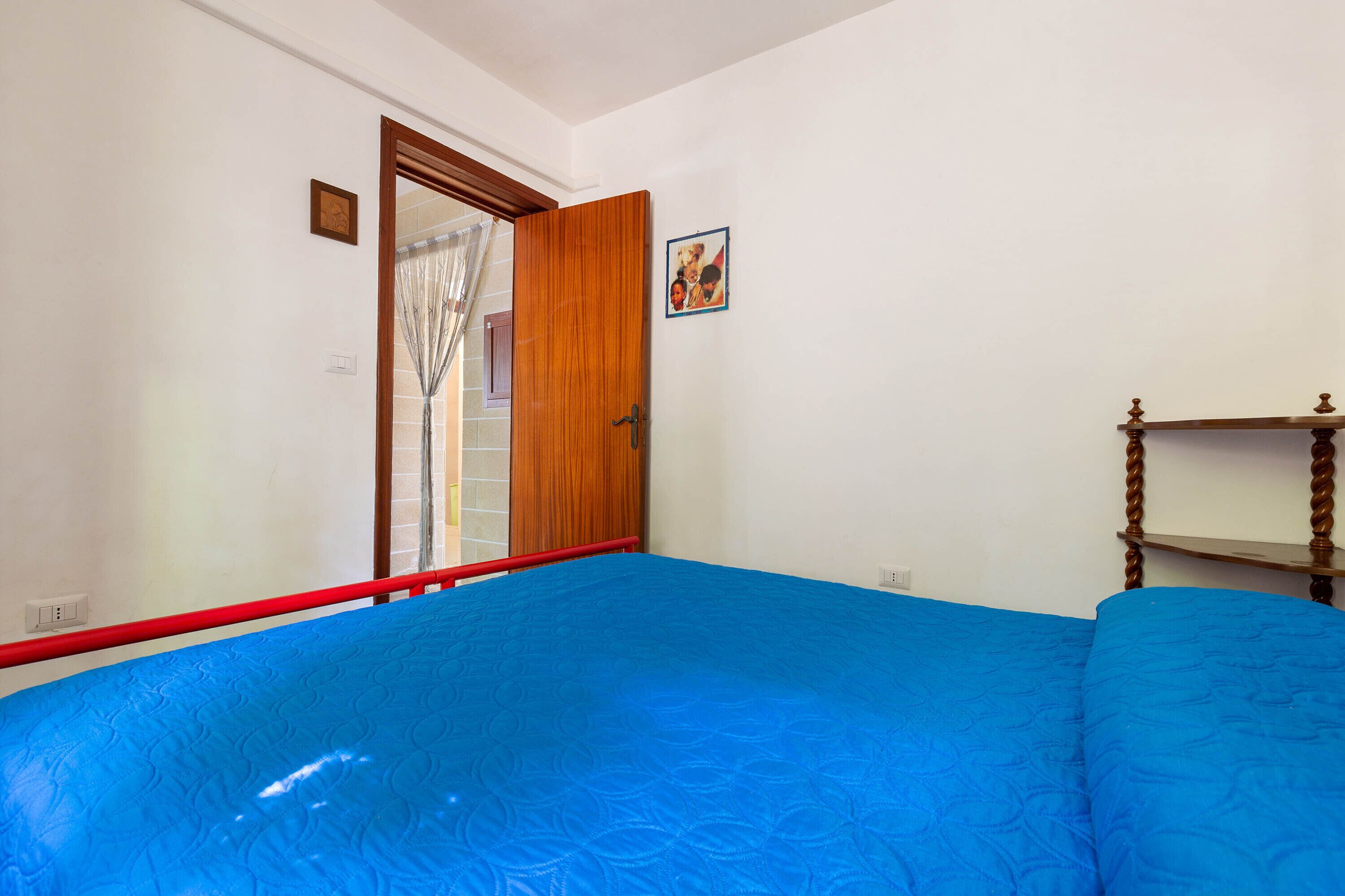 Ein Schlafzimmer mit blauem Bettbezug, einer Holztür, einem kleinen gerahmten Bild und einem Nachttisch aus Holz.
