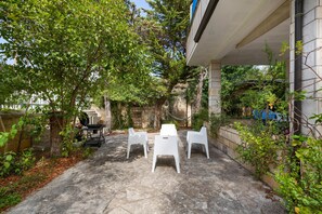 Eine Terrasse mit weißen Plastikstühlen und einem Grill, umgeben von Grünpflanzen und einer Steinmauer.