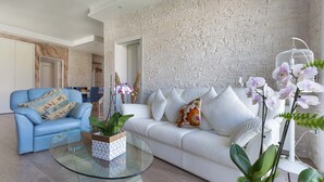 A modern living room with a white sofa, a blue armchair, and a glass coffee table. There are potted plants and a white birdcage with flowers. - Villa Padangi (Gallipoli)