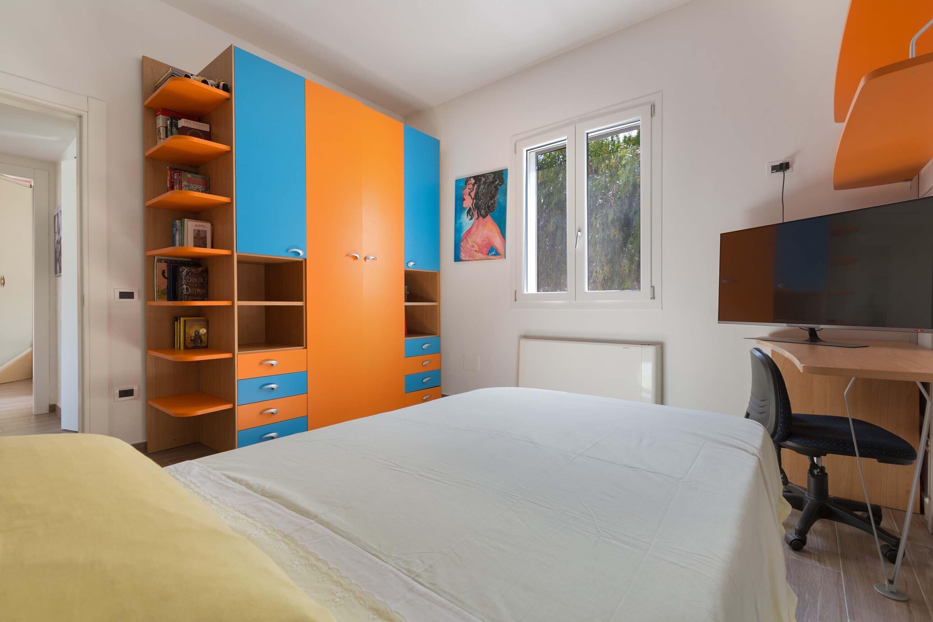 A bedroom with a bed, a desk with a computer, a bookshelf, and a window.
