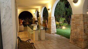 An outdoor patio area with a glass table and chairs, surrounded by stone walls and arches, featuring a small garden and a view of a lit interior space. - 2099 Villa Arkè (Alliste)