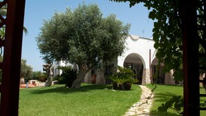 A stone pathway leading to a building with an arched entrance, surrounded by well-maintained grass and a large tree. - 2099 Villa Arkè (Alliste)