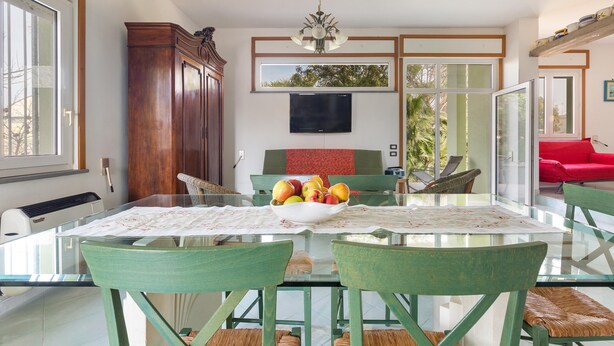 A dining area with a glass table, green chairs, a bowl of fruit, and a television on the wall. - 2143 Villa Corsarina (Nardò)