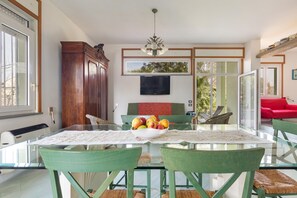 Un coin repas avec une table en verre, des chaises vertes, un saladier rempli de fruits et un téléviseur fixé au mur.