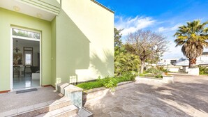 A building with a light green exterior, a tiled entrance, and a paved courtyard with a palm tree and plants. - 2143 Villa Corsarina (Nardò)