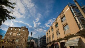 Una calle con una mezcla de edificios antiguos de piedra y arquitectura moderna, incluyendo un edificio con balcón y uno con toldo a rayas.