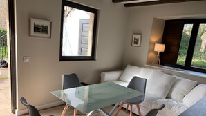 Una sala de estar moderna con una mesa de vidrio, dos sillas, un sofá blanco y una ventana con vista a la vegetación.