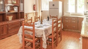 Un coin repas avec une table en bois, dressée pour quatre personnes, entourée de chaises. On aperçoit un réfrigérateur blanc et une fenêtre avec des stores à l’arrière-plan.