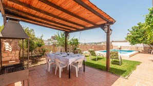 Une terrasse couverte avec une table dressée pour les repas, un barbecue et une piscine en arrière-plan.
