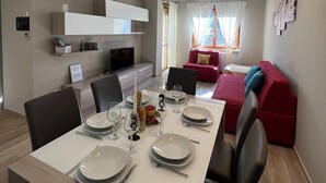 Ein modernes Esszimmer mit einem langen Tisch, gedeckt für vier Personen, einer roten Couch im Hintergrund und einer Küchenzeile mit einer Mikrowelle und einem Fernseher.