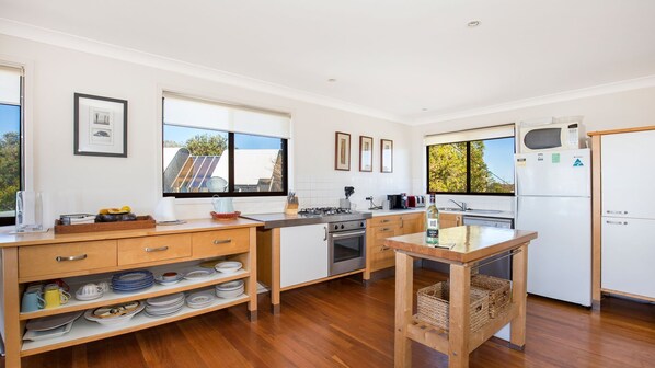 A modern kitchen with wooden cabinets, a gas stove, a microwave, and a refrigerator. There are windows providing natural light and a view of the outside. - Endless Summer - Pet Friendly Absolute Beachfront (Culburra Beach)