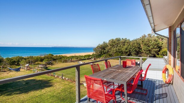 Un balcón con mesa de madera y sillas rojas, con vista a una playa y el océano.
