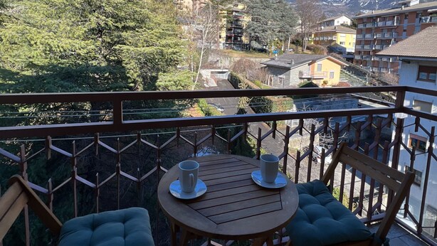 Een balkon met een houten tafel en twee comfortabele stoelen, met uitzicht op een bergachtig landschap met gebouwen.