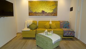 Een gezellige woonkamer met een zithoek, een groen poefje en een kleurrijke plaid.