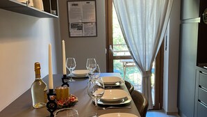 Een eetruimte met een lange tafel voor zes personen, compleet met borden, glazen en bestek. Er staan kaarsen en een fles op tafel. De kamer heeft een modern design, met een groot raam en gordijnen.
