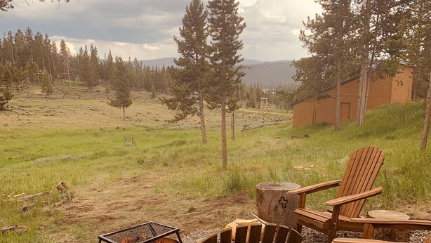 An outdoor seating area with wooden chairs and a fire pit, surrounded by a natural landscape with trees and a distant building.