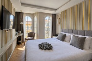 A hotel room with a large bed, a television, a desk with a chair, and a view of buildings through large windows. - Serai Hotel (Istanbul)
