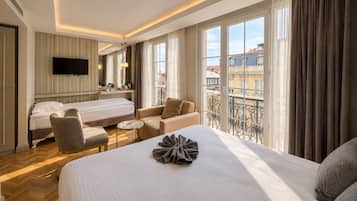 Habitación de hotel con cama, sofá, sillón, mesita, televisión y balcón con vista a edificios.