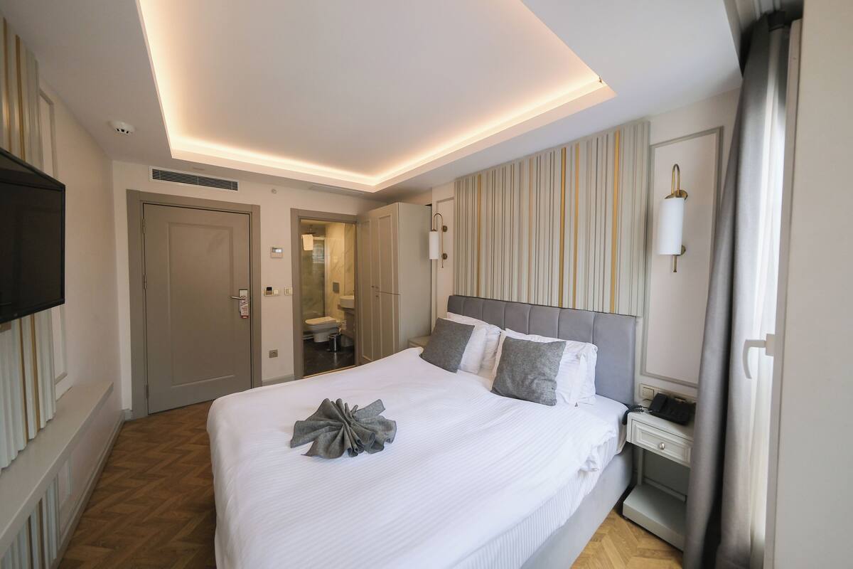 A hotel room with a neatly made bed, a bedside table with a lamp, a flat-screen TV mounted on the wall, and a bathroom visible through an open door.