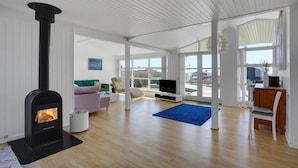 Ein geräumiges Wohnzimmer mit Kaminofen, Fernseher und einem großen Fenster mit Blick ins Freie.