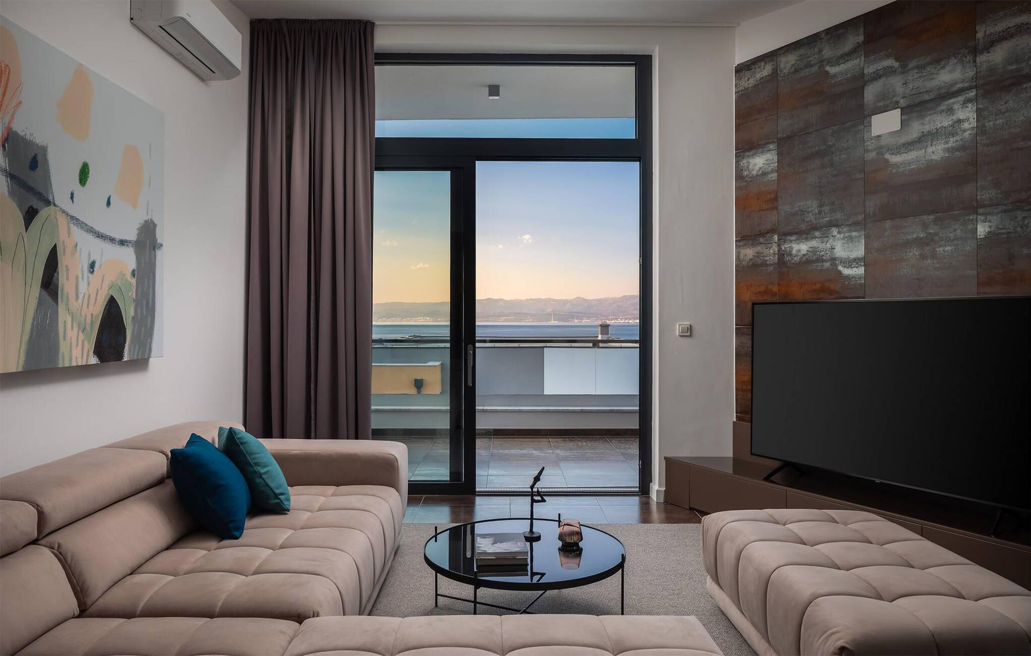 Una sala de estar moderna con un sofá, una mesa de centro y un televisor de pantalla plana grande. Se ve una ventana grande con vistas a la ciudad y a las montañas.