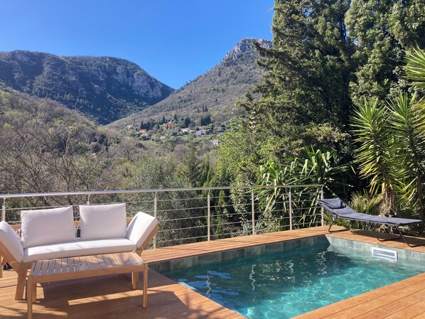 Una terraza de madera con piscina, sillas de descanso blancas y vistas a montañas y árboles.