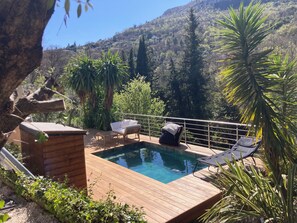 Eine Holzterrasse mit Pool, Liegestühlen und einer kleinen Holzhütte, umgeben von Grünflächen und Hügeln.