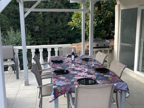 Un área de comedor al aire libre cubierta con una mesa preparada para cuatro personas, rodeada de sillas, y una parrilla al fondo.