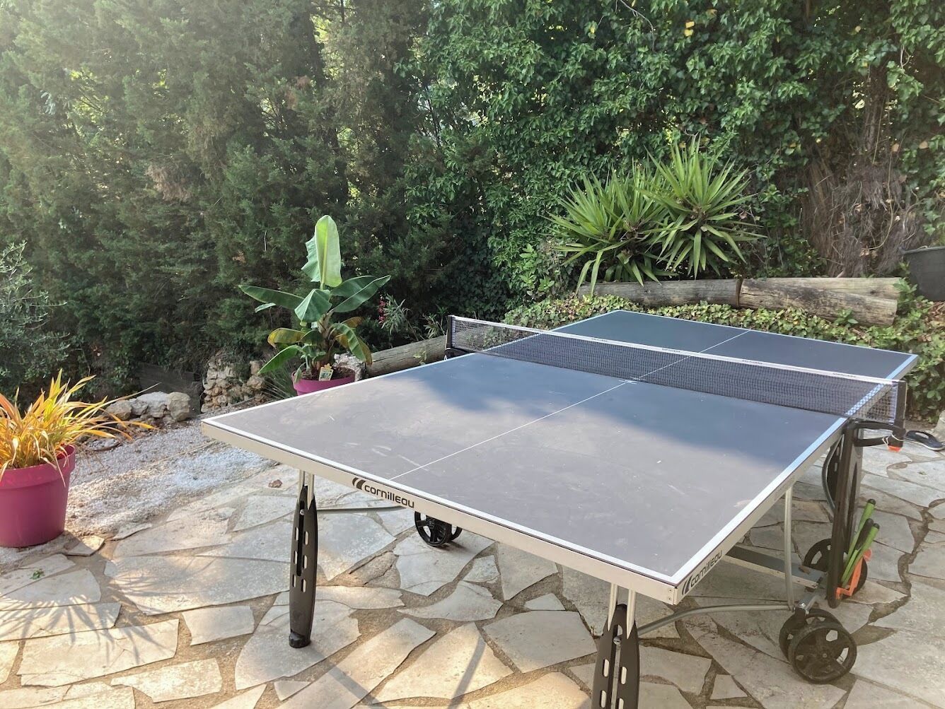 A portable ping pong table with wheels, set on a stone patio surrounded by potted plants.