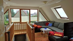 A cozy attic room with a sloped ceiling, large windows, a built-in wooden staircase, a grey sofa with colorful cushions, and a small table with a tablecloth. - Wunderschöne Ferienwohnung (Brandenburg an der Havel)