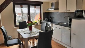 A modern kitchen with a dining area, featuring a table with chairs, a window with lace curtains, and a view of the outside. - Wunderschöne Ferienwohnung (Brandenburg an der Havel)
