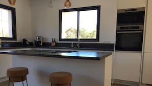 A modern kitchen with a large island, two stools, a built-in oven, and a window providing natural light. - Maison VUE MER 50M DE LA Plage A Pied (Hyères)